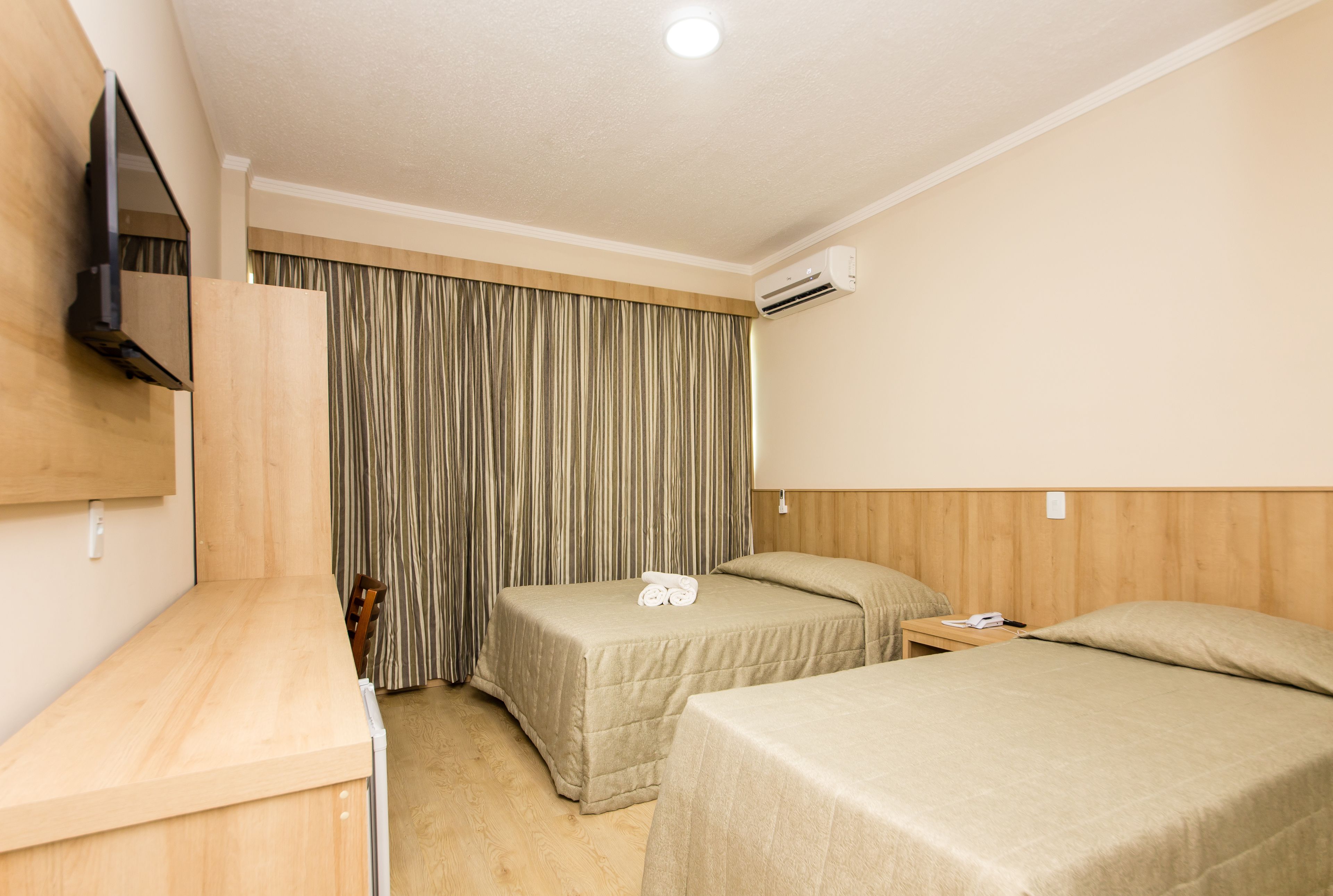 A hotel room with two beds, a wooden desk, a TV mounted on the wall, and an air conditioning unit.