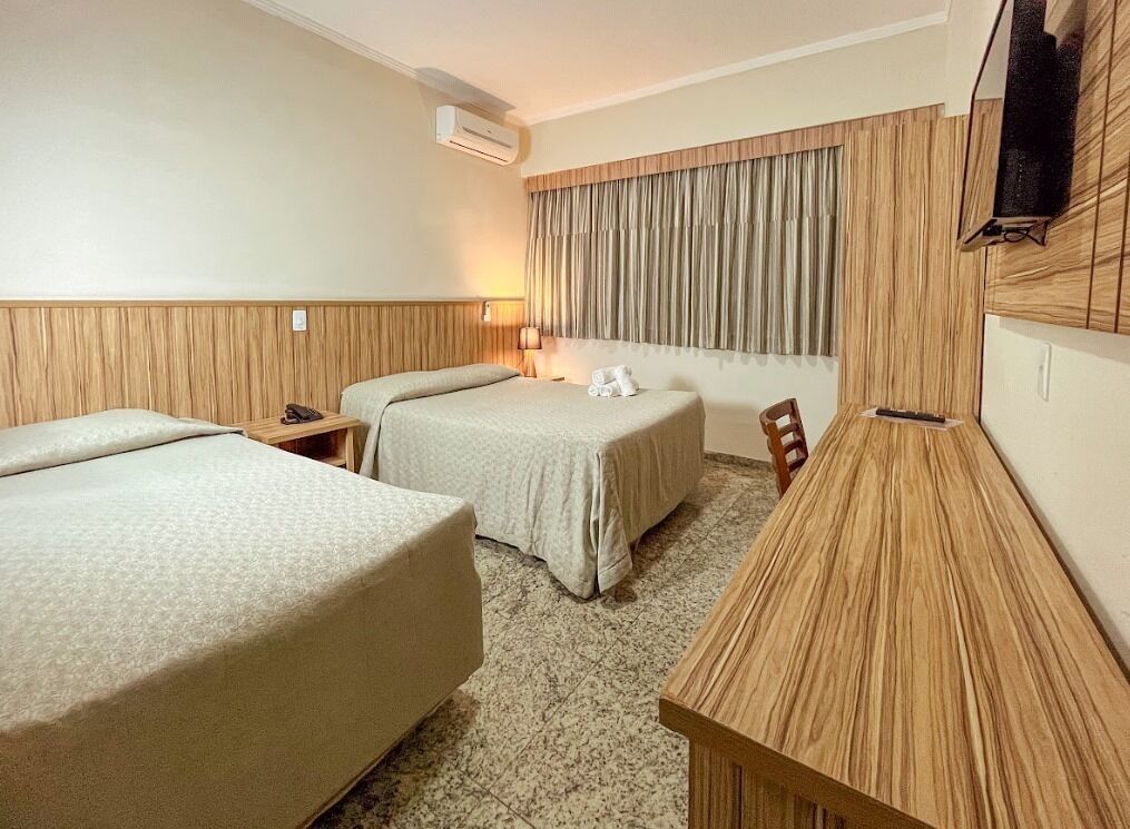A hotel room with two beds, a desk, a chair, a TV, and a window with curtains.