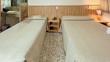 Quarto de hotel com duas camas de solteiro, uma cabeceira de madeira, um criado-mudo de madeira com um telefone, uma escrivaninha com uma luminária e um banheiro visível ao fundo.