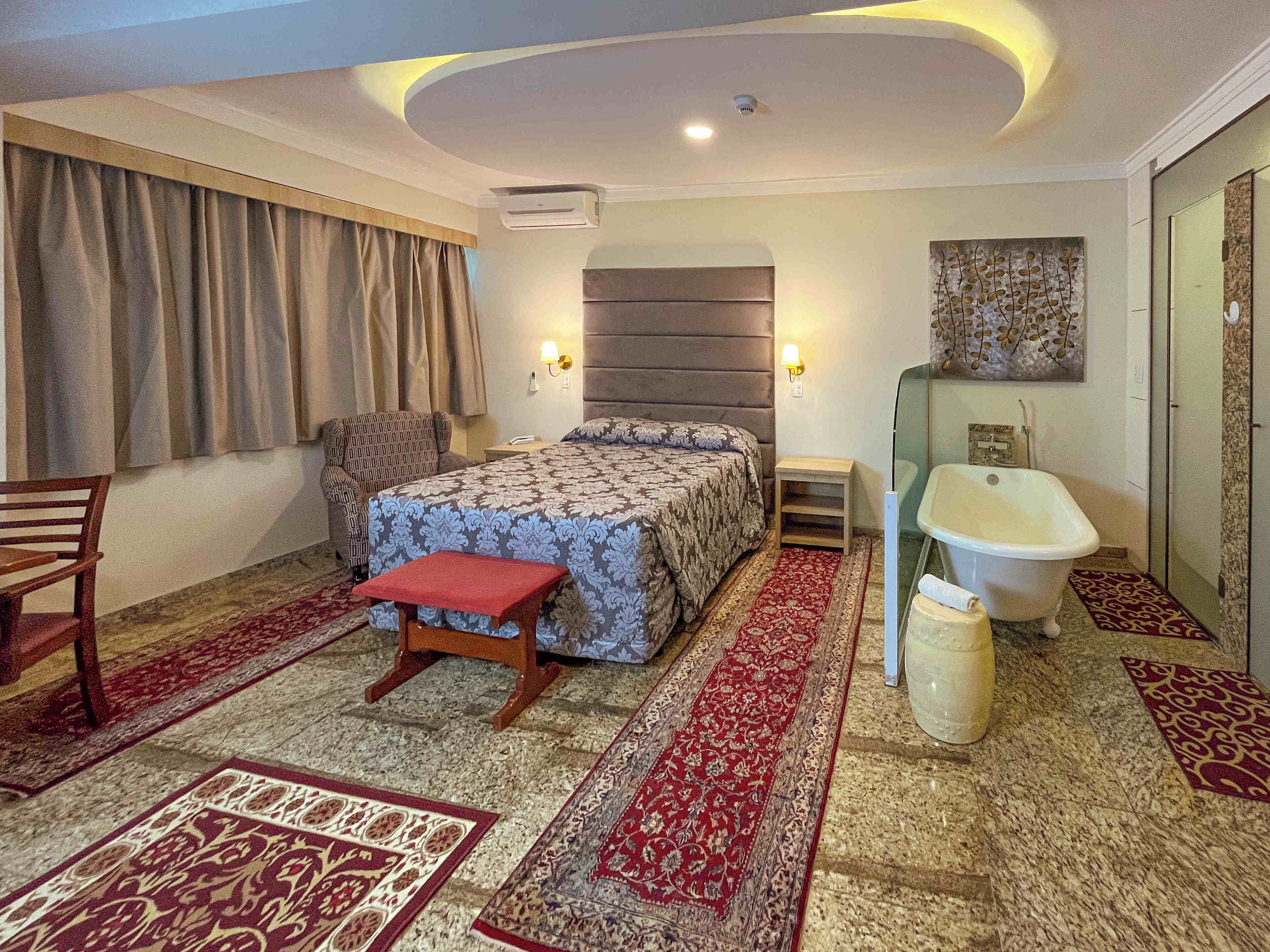 Un dormitorio con cama, una silla, una mesita de noche, aire acondicionado y un baño con bañera y ducha.