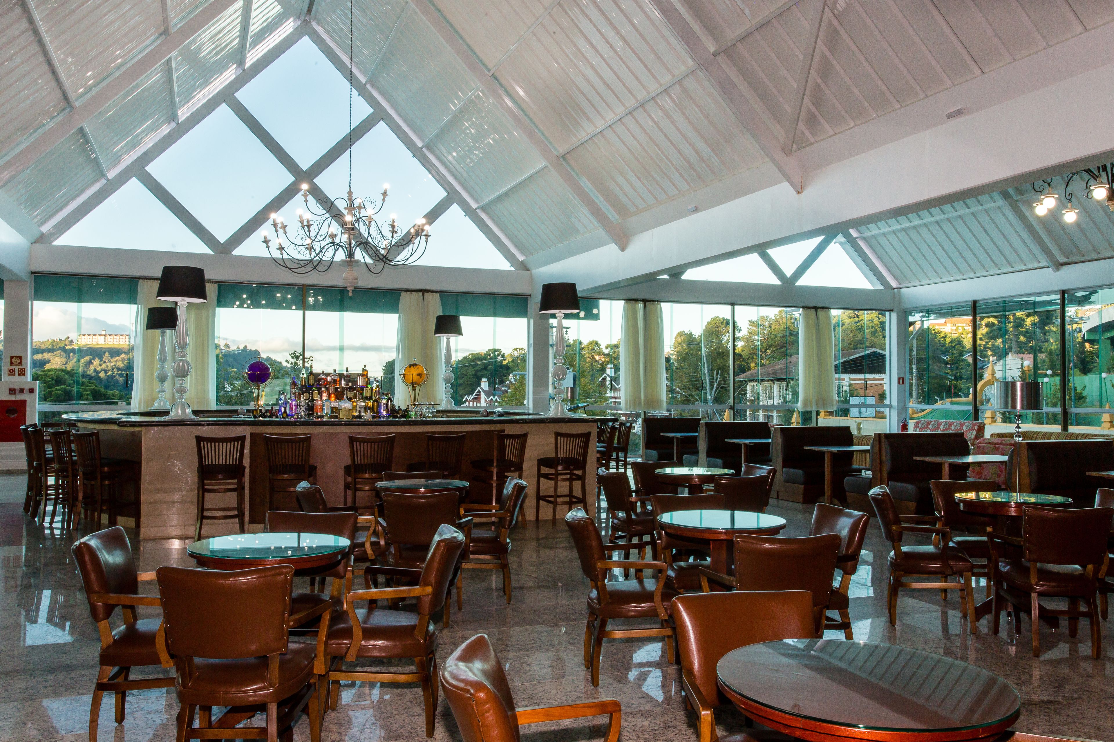 A spacious indoor area with a high ceiling, large windows, and a bar area with chairs and tables.