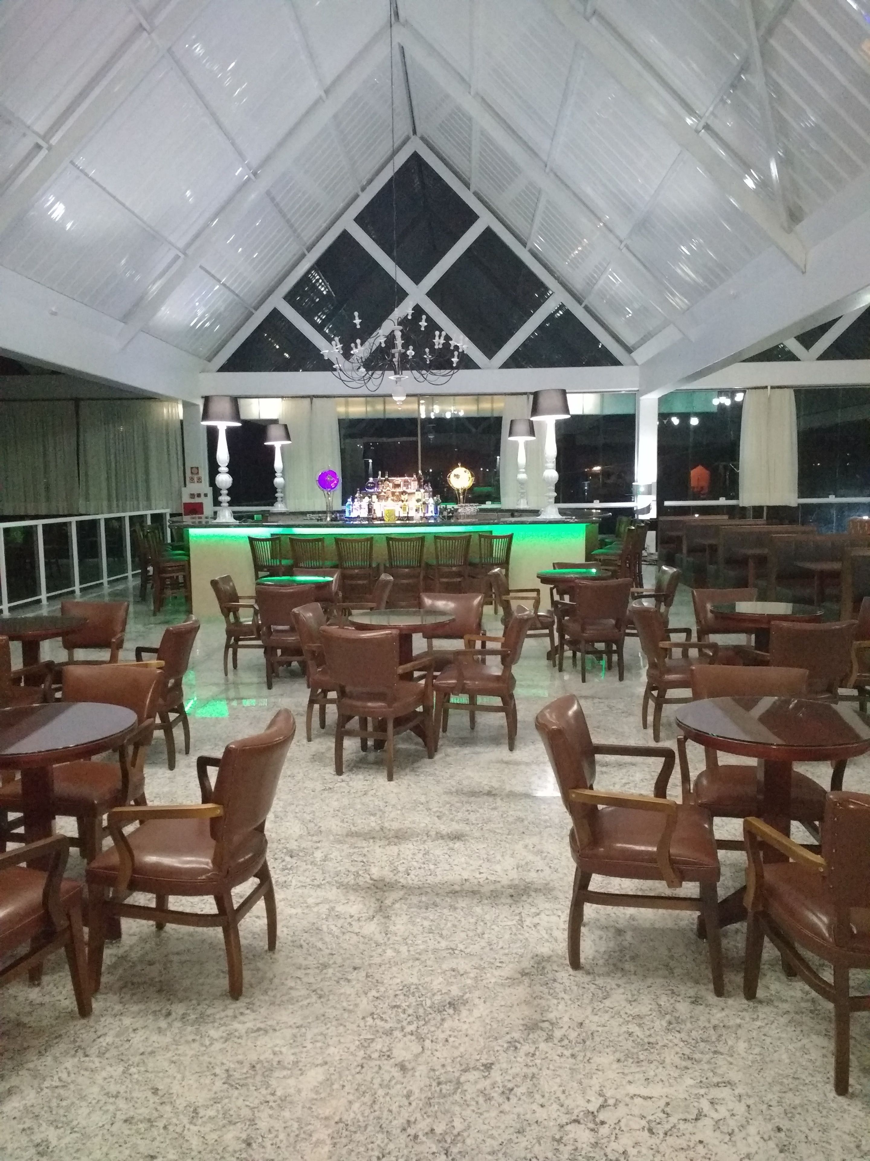 Ruime overdekte eetzône met een hoge plafond en een podium aan de voorzijde. Tafels en stoelen staan in rijen opgesteld, en er is een bar met een verlichte toog.