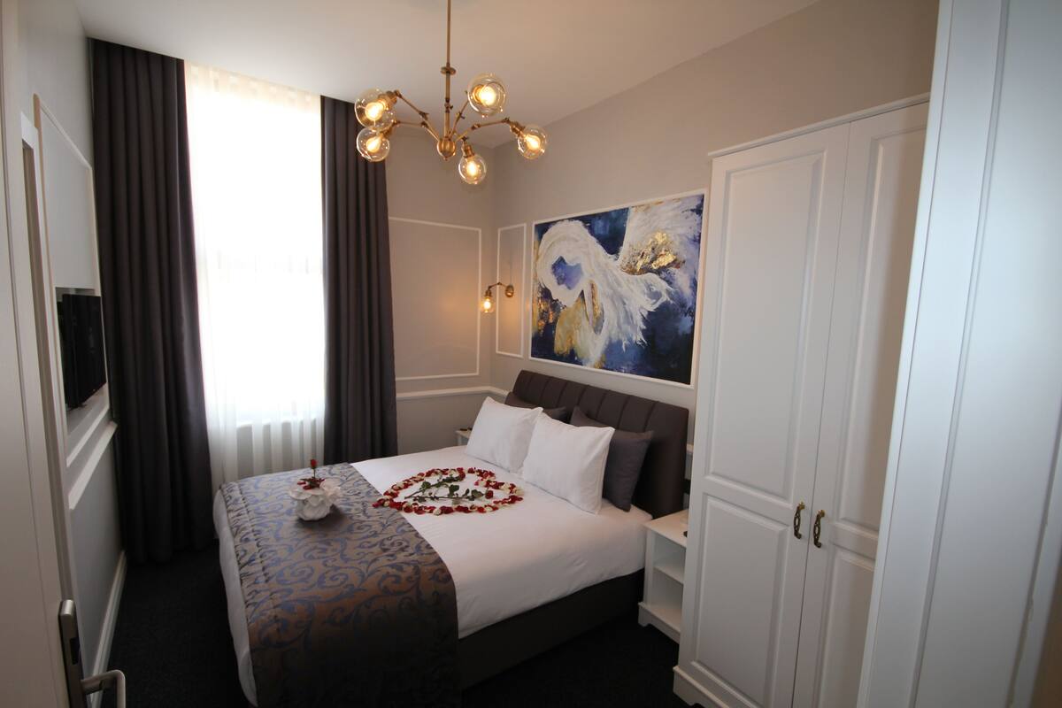 A hotel room with a bed, a chandelier, a painting on the wall, and a wardrobe.