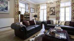 A living room with a leather sofa set, a coffee table with books and glasses, floral curtains, and a chandelier. - Town House at Brecon (Brecon)