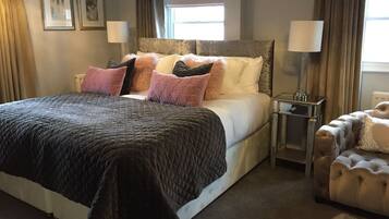 A bedroom with a large bed, a nightstand, a lamp, and a tufted armchair.