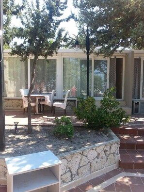 Un patio al aire libre con un banco de piedra, una mesa con sillas y un pequeño jardín con arbustos.