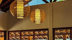 Een restaurant met houten tafels en stoelen, grote ramen met uitzicht op zee, en hangende rieten lampen.