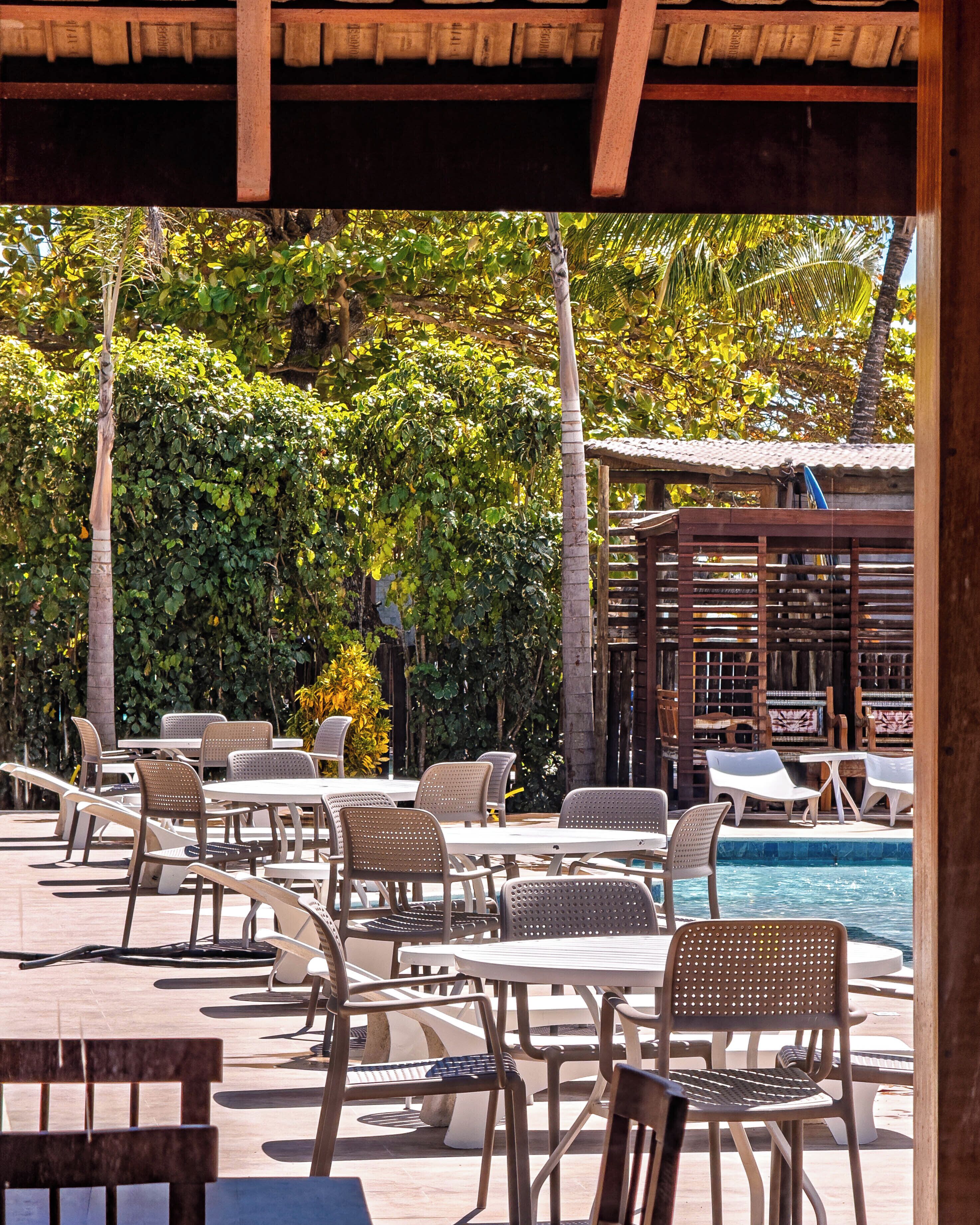 An outdoor pool area with lounge chairs and tables, surrounded by lush greenery and a wooden pergola.
