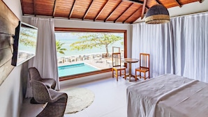 Een kamer met houten plafond, een bed, een televisie, uitzicht op een zwembad en strand, en een stoel bij het raam.