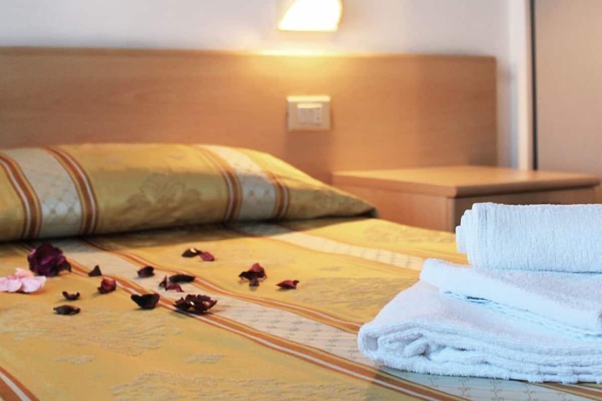 A neatly made bed with a pillow, a folded towel, and scattered rose petals.