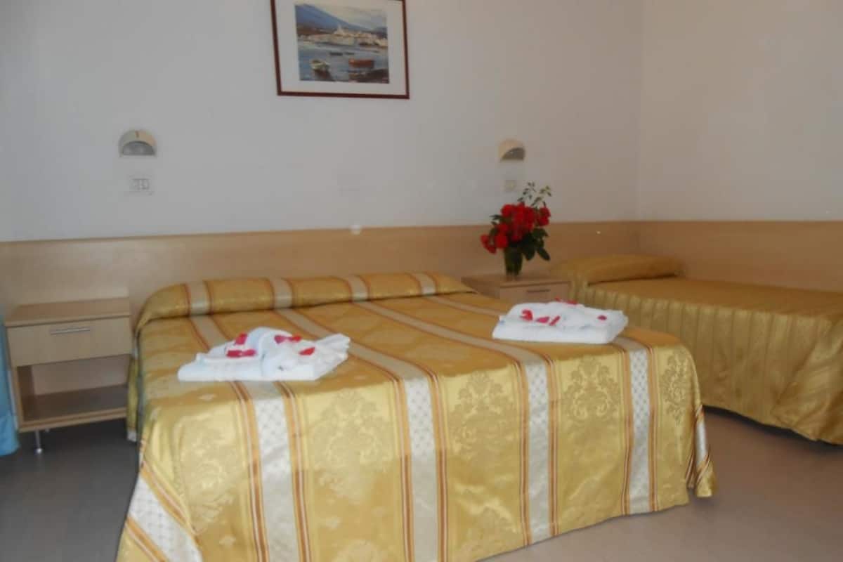 A hotel room with two beds, each with a yellow bedspread and white pillows. There is a small table with a vase of red flowers and a framed picture on the wall.