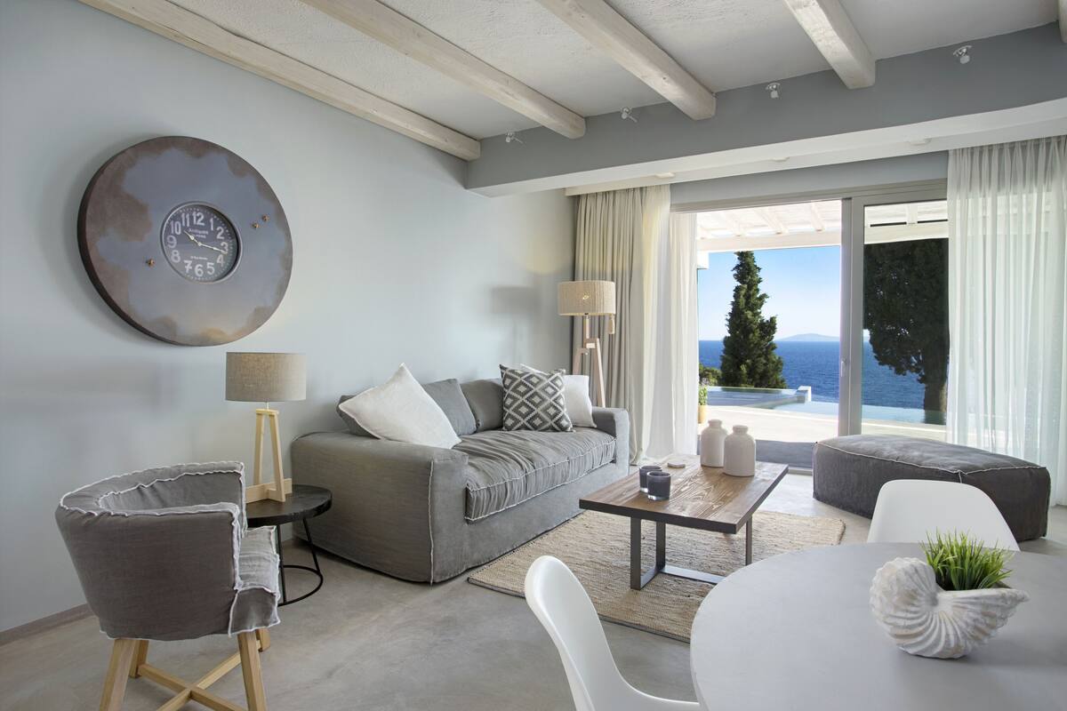 Een moderne woonkamer met een grijze bank, een ronde salontafel en uitzicht op zee door grote ramen.