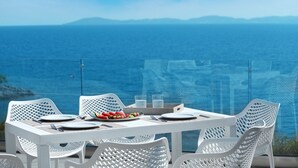 Une table dressée pour deux personnes, avec des chaises blanches, une assiette de fruits et un verre d’eau, offrant une vue sur la côte.