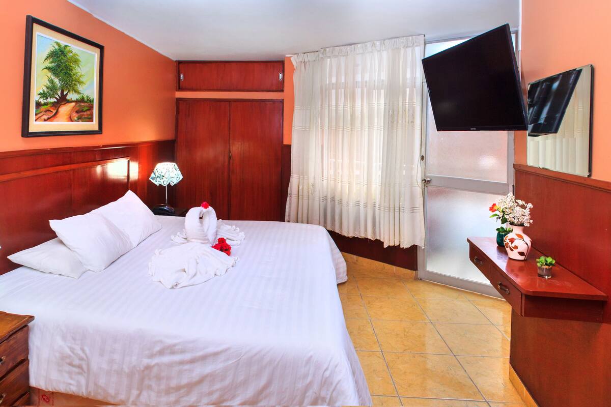 A hotel room with a bed, a nightstand, a TV, a window with curtains, and a painting on the wall.