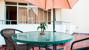 A round glass table with a small plant centerpiece, surrounded by wicker chairs, under a large umbrella, on a tiled patio with a building in the background.