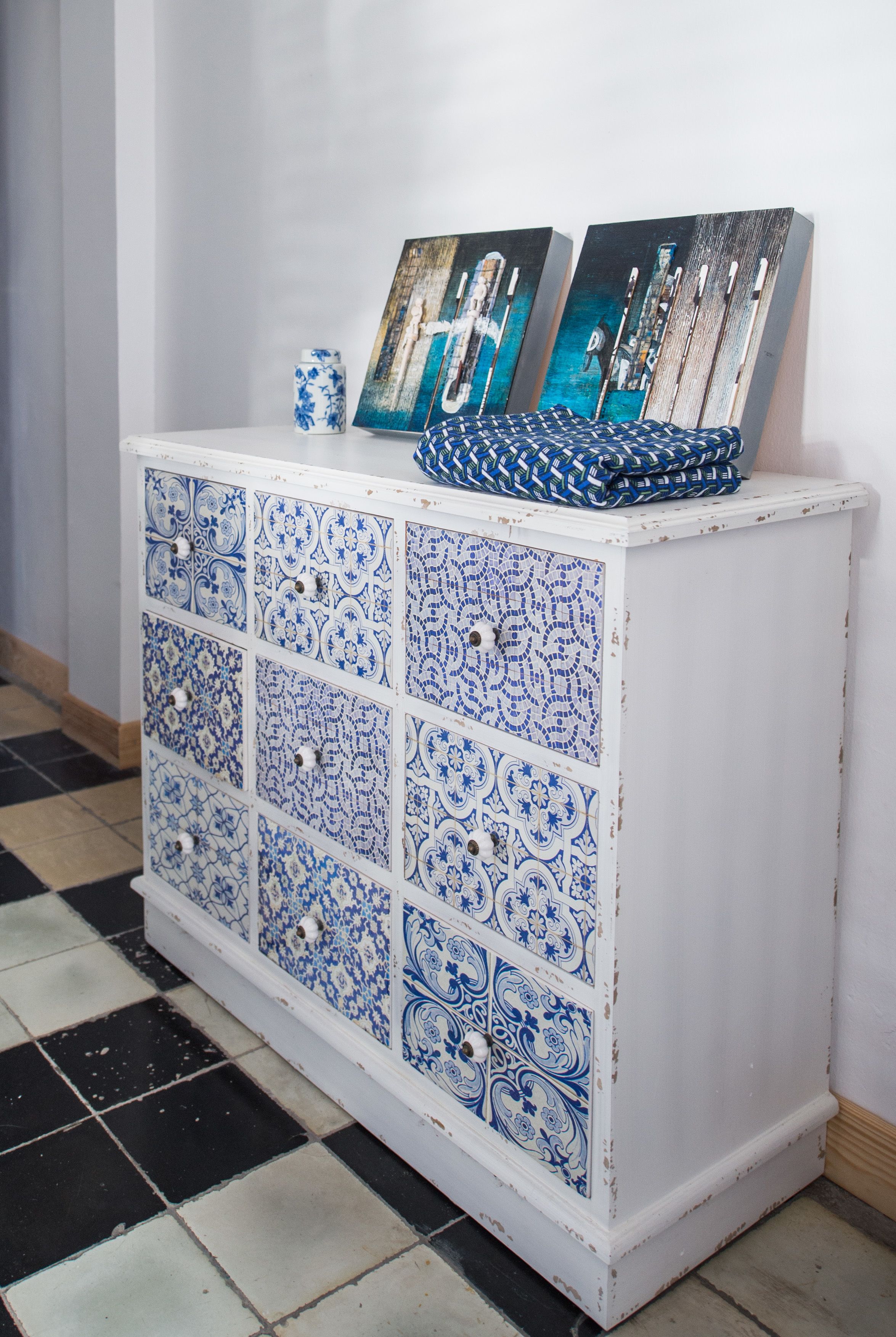 A white cabinet with blue and white patterned tiles and two framed pictures on top.