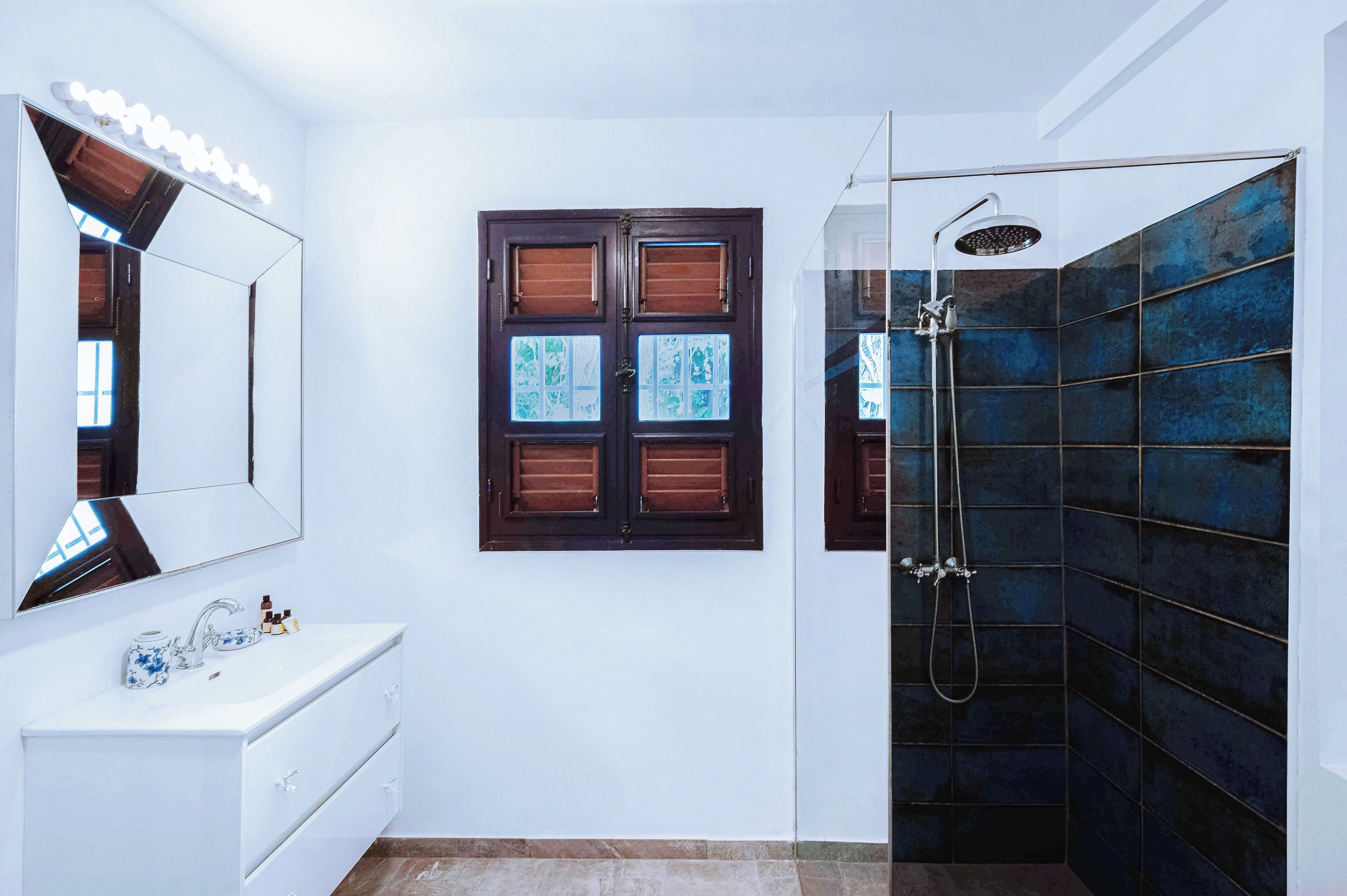 A bathroom with a white vanity, a large mirror, a shower with dark blue tiles, and a window with wooden shutters.