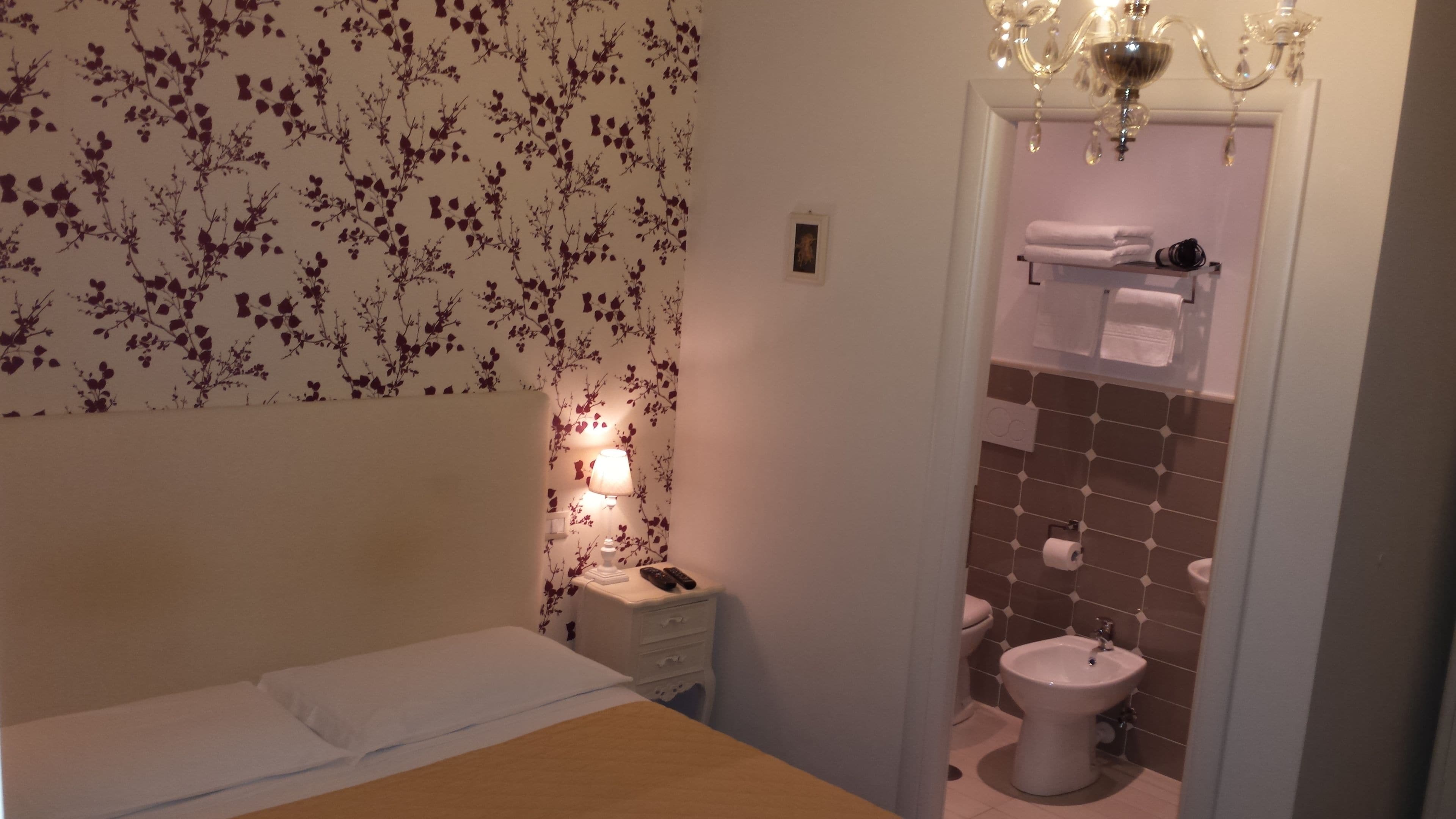 A bedroom with a bed, a bedside table with a lamp, and a bathroom with a toilet, sink, and towel rack.