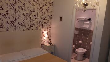A bedroom with a bed, a bedside table with a lamp, and a bathroom with a toilet, sink, and towel rack.