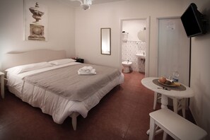 A hotel room with a bed, bedside table, a small round table with a fruit bowl, a bench, a TV mounted on the wall, and a bathroom visible through an open door. - Domus Flaviae (Rome)