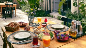 A wooden table set with breakfast items, including bread, fruit, and a vase of flowers, in a courtyard with stone walls and greenery. - Manastir Alacati Hotel (Cesme)