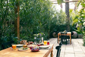 Una mesa de madera dispuesta con diversos platos, bebidas y un jarrón de flores en un jardín exuberante.