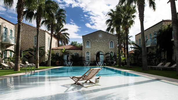 A swimming pool with lounge chairs and palm trees, surrounded by stone buildings with balconies. - Manastir Alacati Hotel (Cesme)