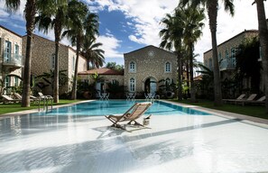 Una piscina con sillas de descanso y palmeras, rodeada de edificios de piedra con balcones.