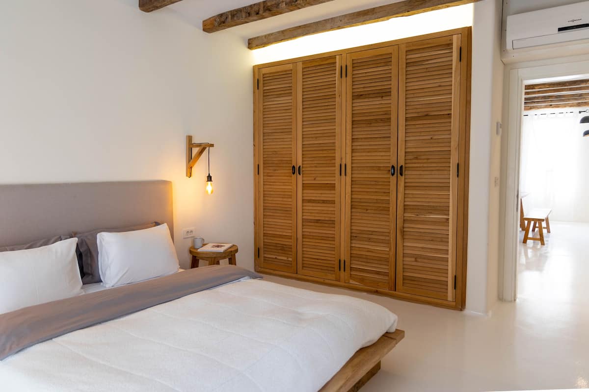 A bedroom with a large bed, wooden wardrobe, bedside table, and a small table with a lamp.
