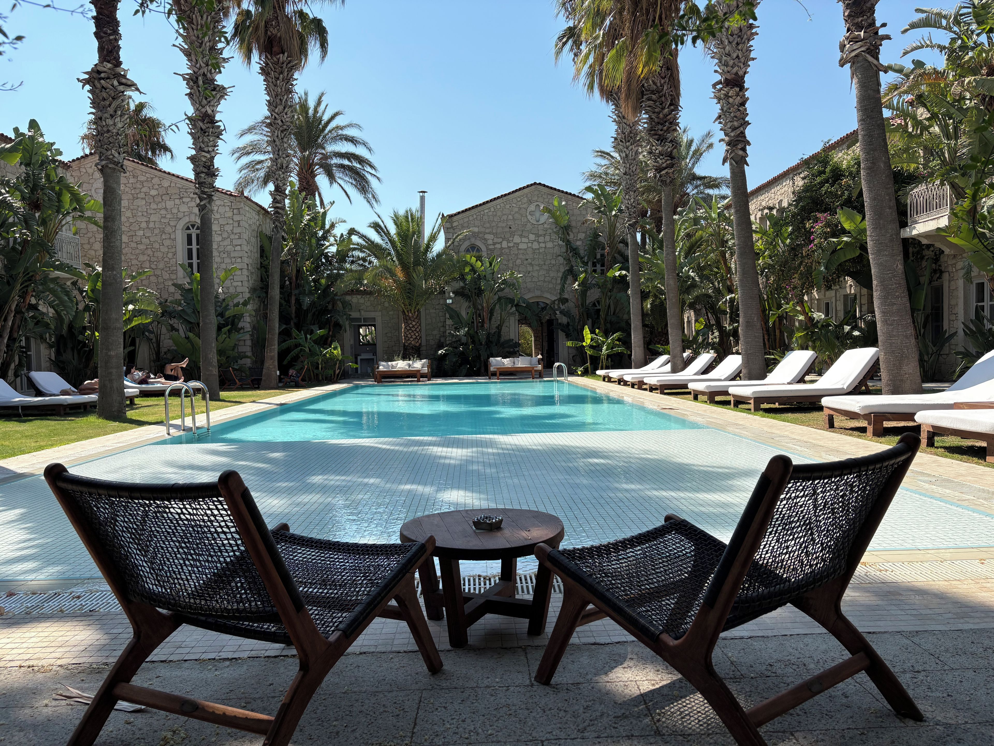 A swimming pool with lounge chairs and a table, surrounded by palm trees and stone buildings.