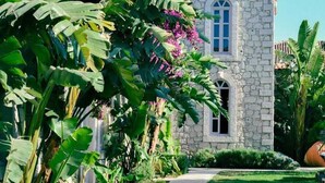 A stone building with a stone facade, a paved pathway, and lush tropical vegetation including banana trees and bougainvillea flowers. - Manastir Alacati Hotel (Cesme)