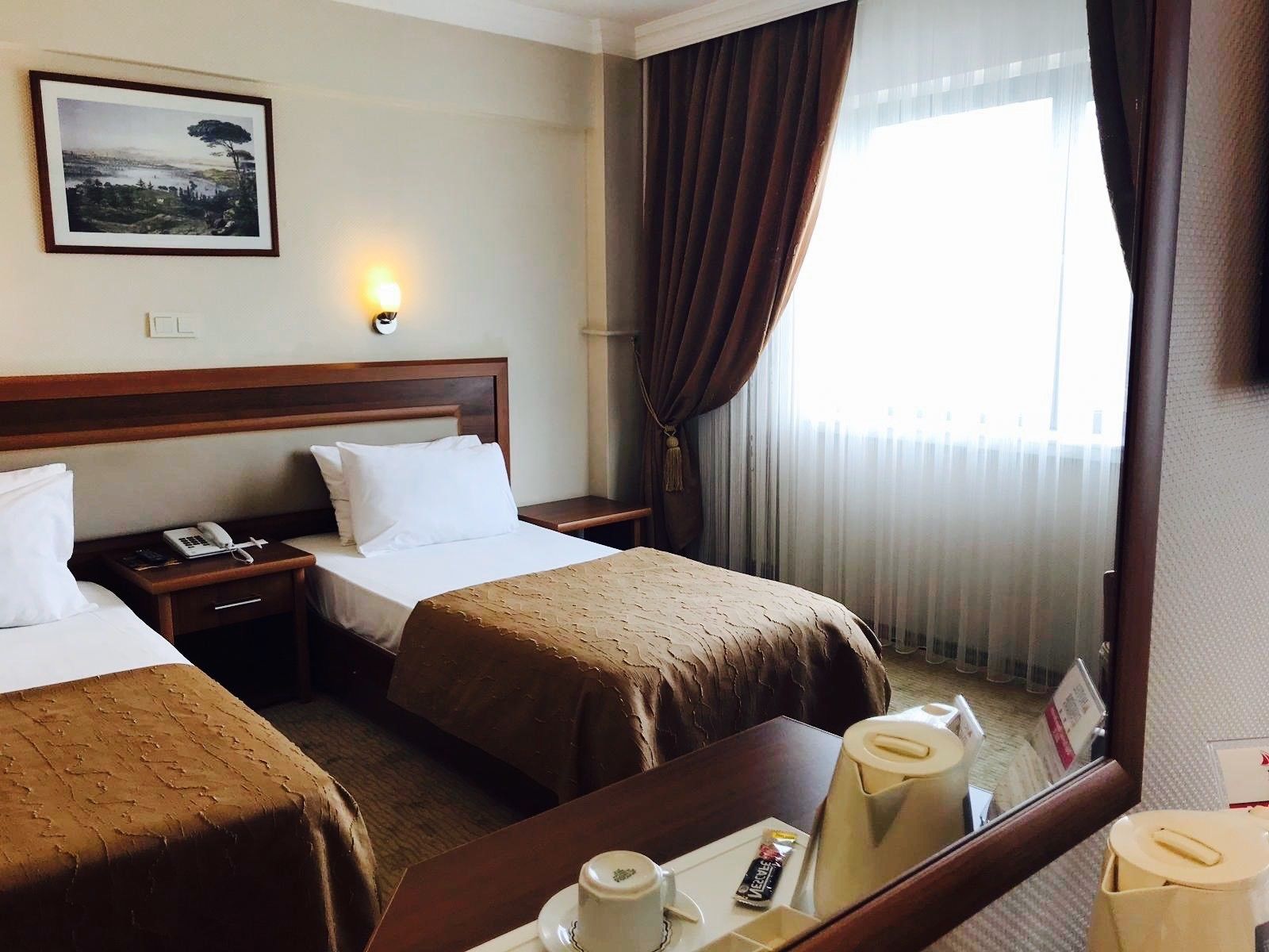 A hotel room with two beds, a nightstand with a phone, a bedside table with a lamp, a coffee maker, and a tray with a cup and a snack.