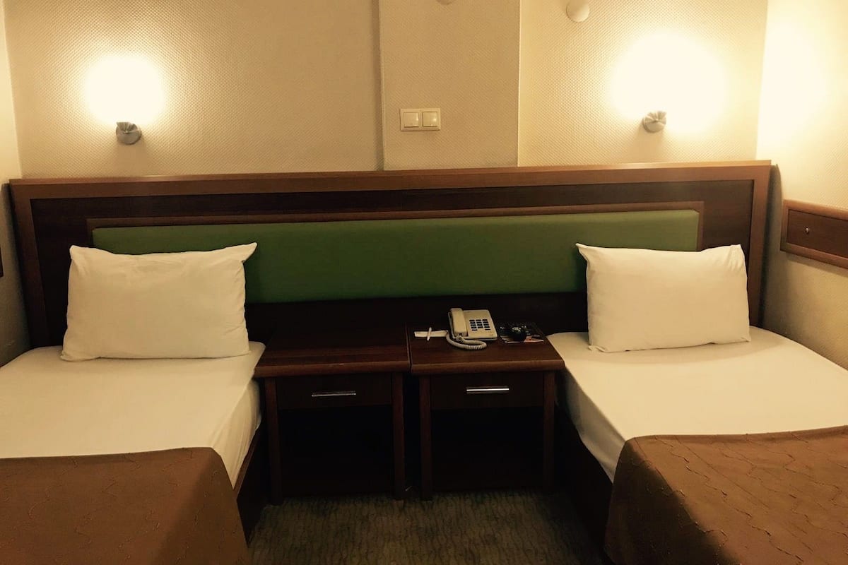 A hotel room with two single beds, a wooden headboard, a bedside table with a telephone, and wall-mounted light fixtures.