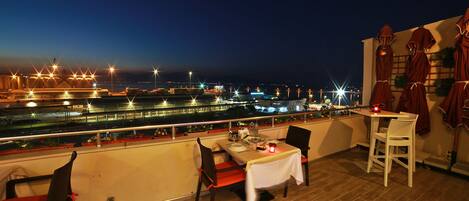 A rooftop dining area with a table set for two, overlooking a cityscape at night.
