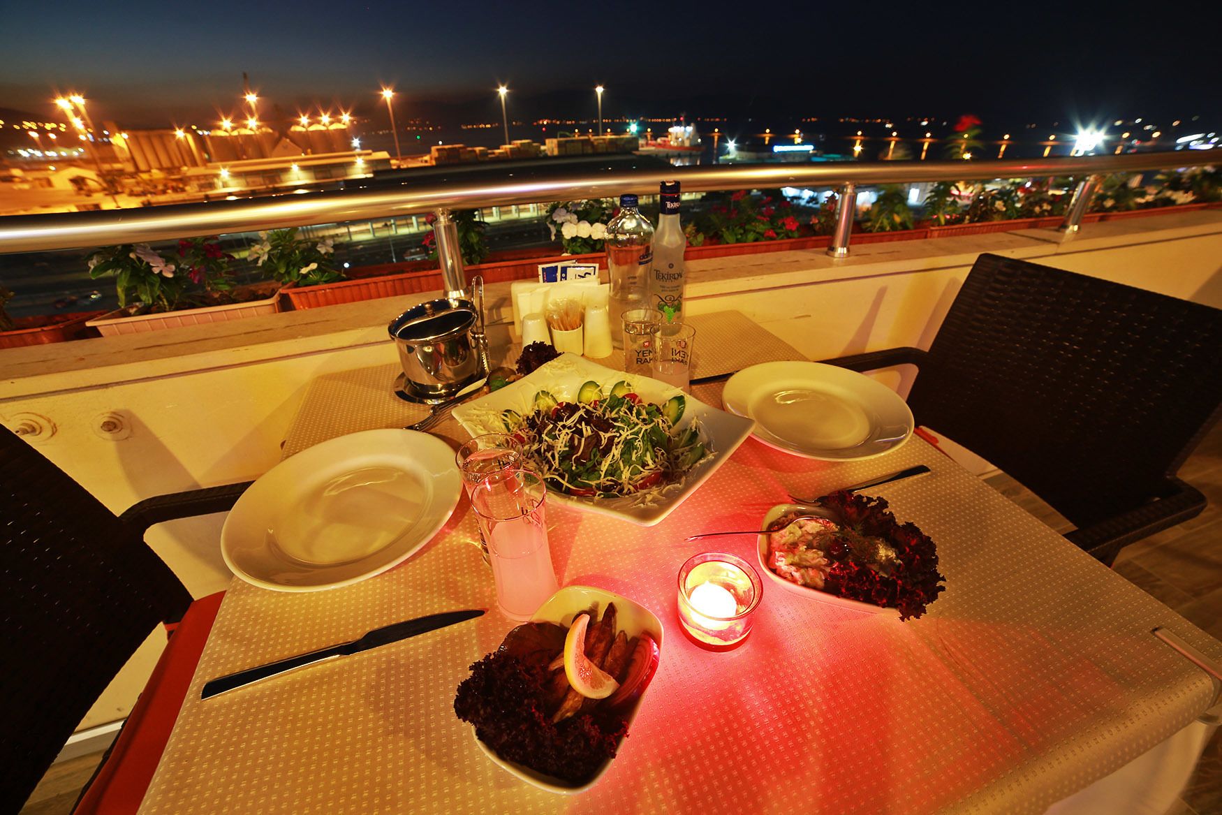 A table set for two with plates, a salad, and a bottle of liquor on a balcony overlooking a cityscape at night.