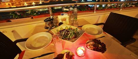 A table set for two with plates, a salad, and a bottle of liquor on a balcony overlooking a cityscape at night.