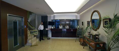 A hotel lobby with a reception desk, a receptionist, a staircase, and a large mirror on the wall.
