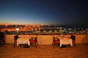 A rooftop dining area with tables set for two, overlooking a cityscape at dusk. - Hotel Villa Marina (Bandirma)