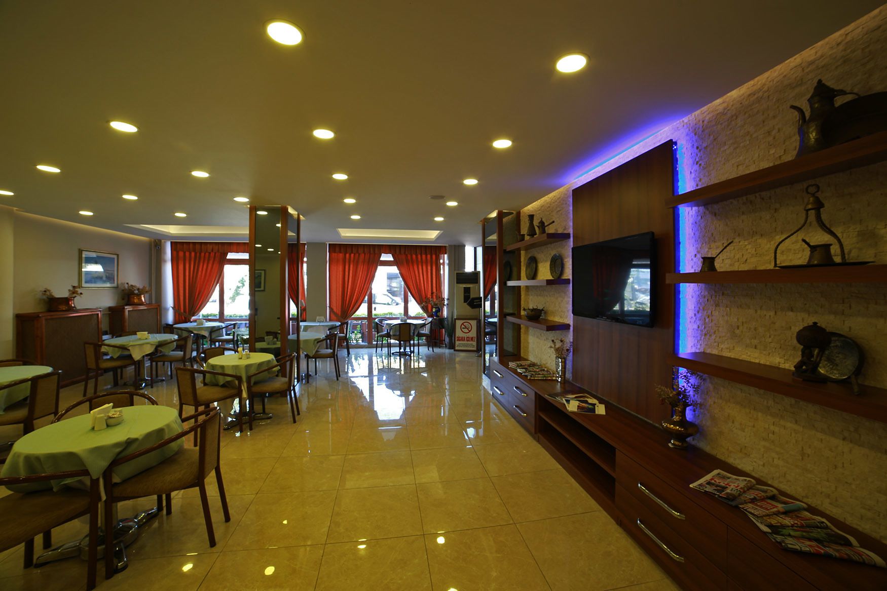 A hotel dining area with multiple tables and chairs, a reception desk, and a wall-mounted television.