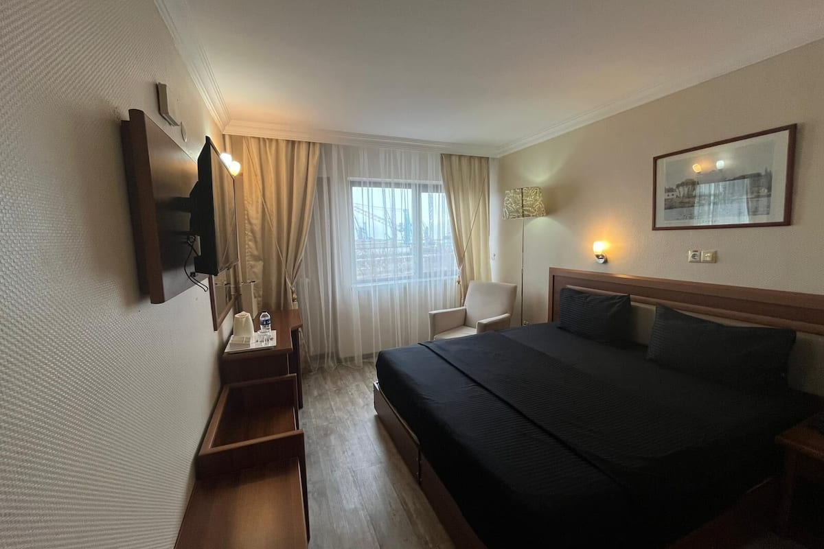 A hotel room with a bed, a desk, a chair, a TV, a window with curtains, and a wall-mounted lamp.