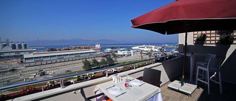 A balcony with a table set for dining, overlooking a port with cargo ships and a clear blue sky.