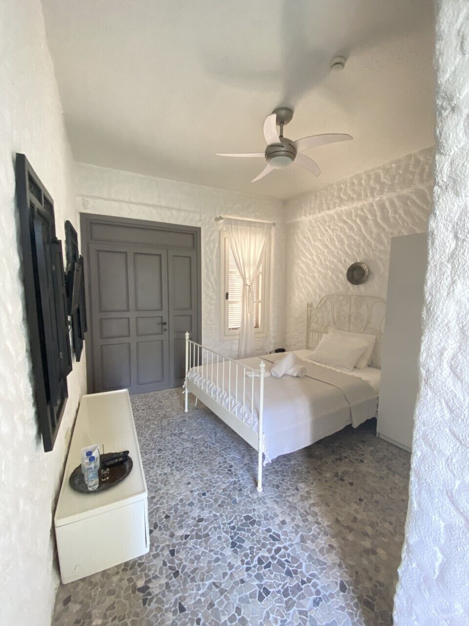 A bedroom with a white bed, a ceiling fan, a door, and a small white table with a bottle and remote.