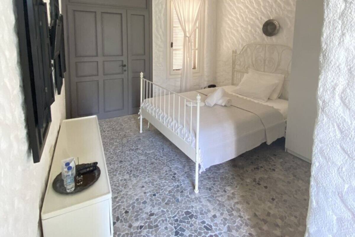 Un dormitorio con cama blanca, ventilador de techo, una puerta y una pequeña mesa blanca con una botella y un control remoto.