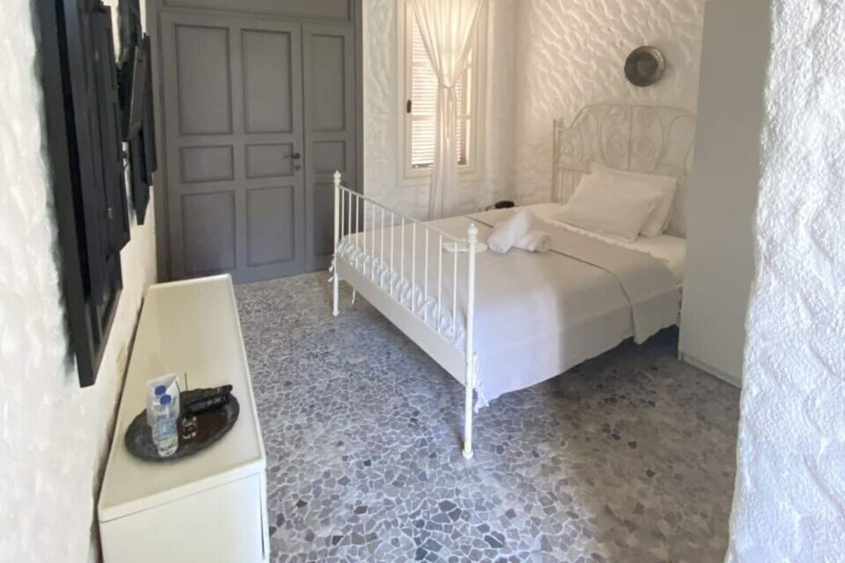 A bedroom with a white bed, a ceiling fan, a door, and a small white table with a bottle and remote.