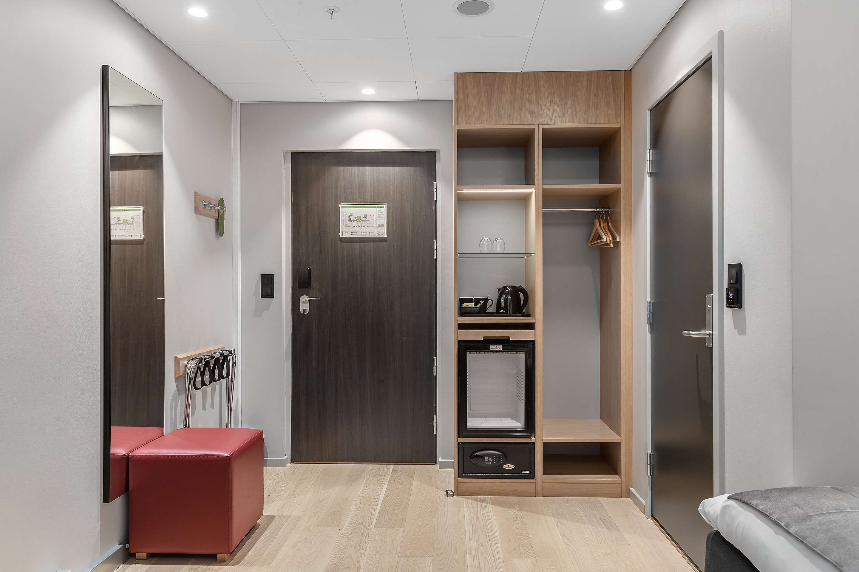A modern hotel room with a wooden wardrobe, a red bench, a small refrigerator, and a bed.