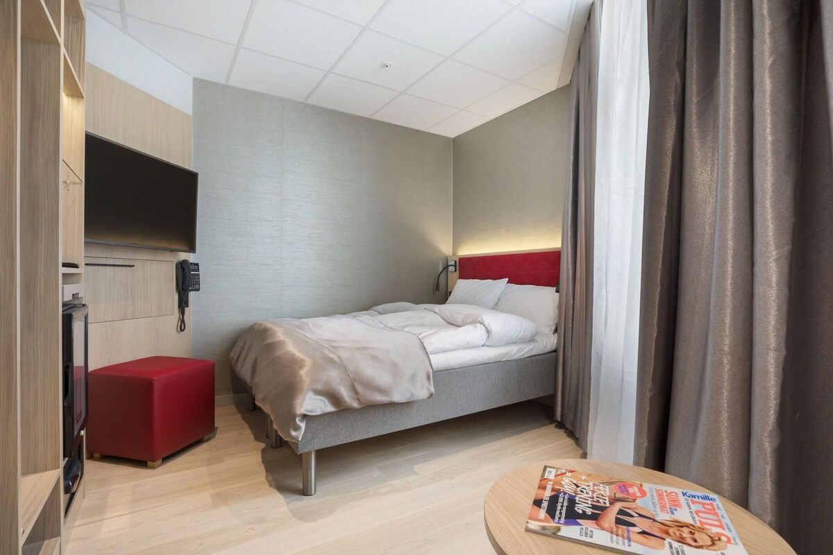 A hotel room with a bed, a red ottoman, a round table with a magazine, and a TV mounted on the wall.