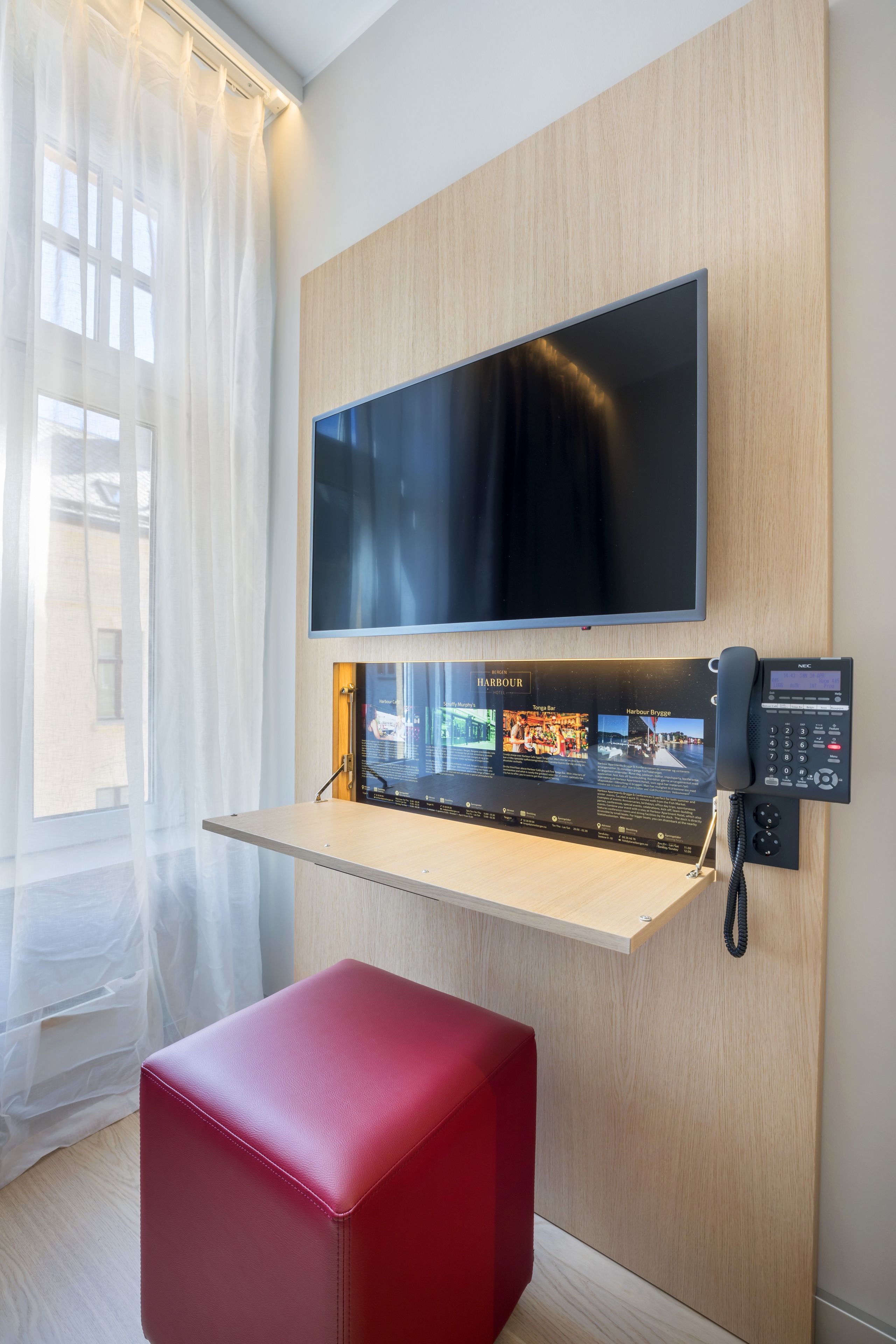Habitación de hotel con una televisión de pantalla plana montada, un escritorio con teléfono y un otoman rojo.
