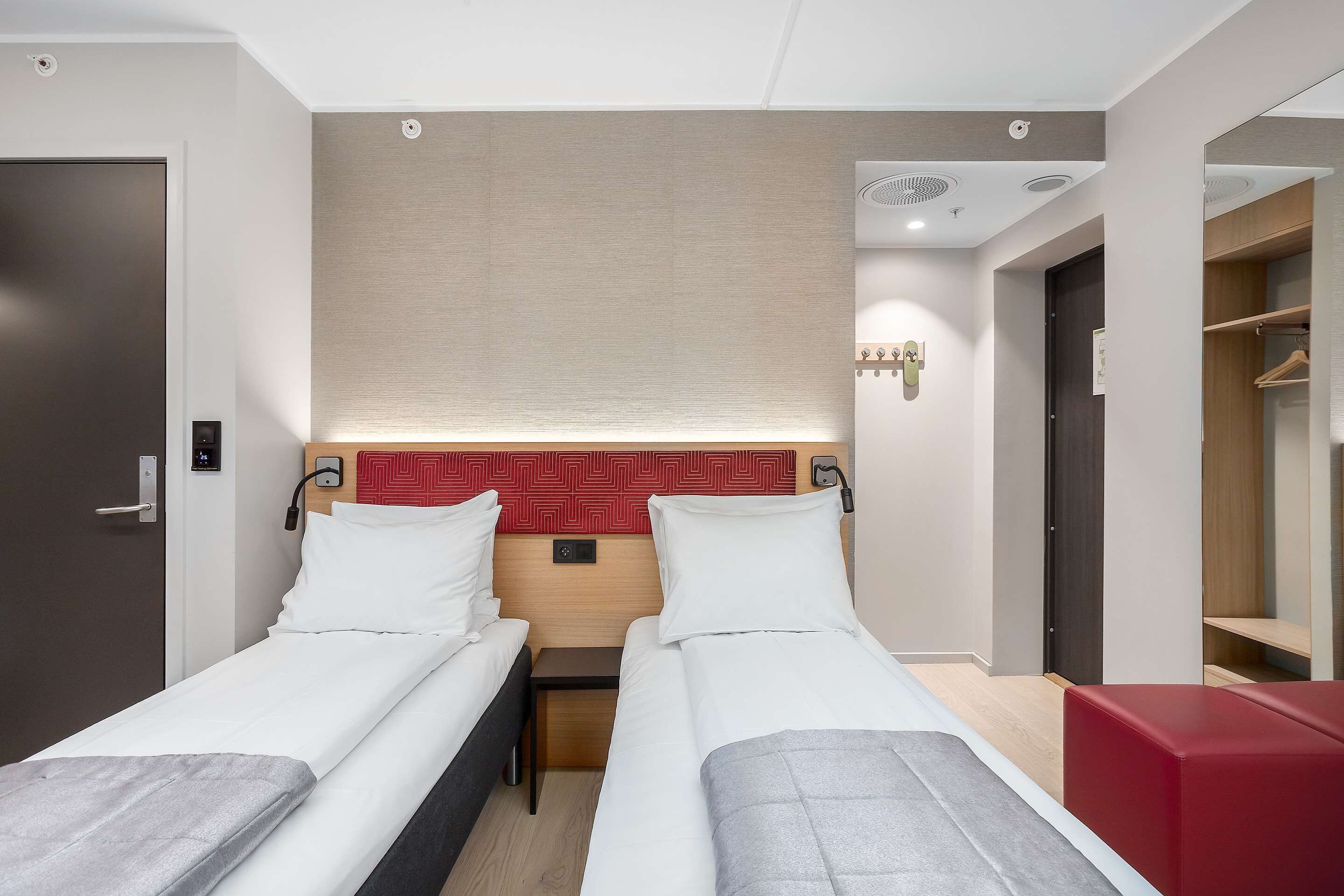 Habitación de hotel con dos camas individuales, una mesita de noche pequeña, cabecero rojo, armario y zona de estar en rojo.