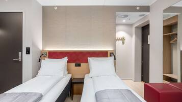 Habitación de hotel con dos camas individuales, una mesita de noche pequeña, cabecero rojo, armario y zona de estar en rojo.