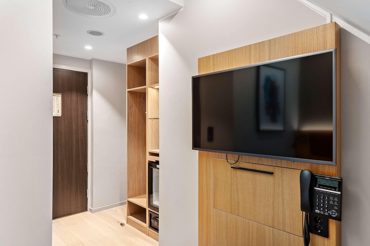 A modern hotel room with a built-in TV, a telephone, and a wooden wardrobe.